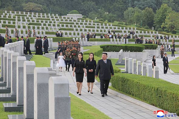 A solemn procession of individuals, dressed in black, walks through a meticulously maintained cemetery. The graves are aligned i